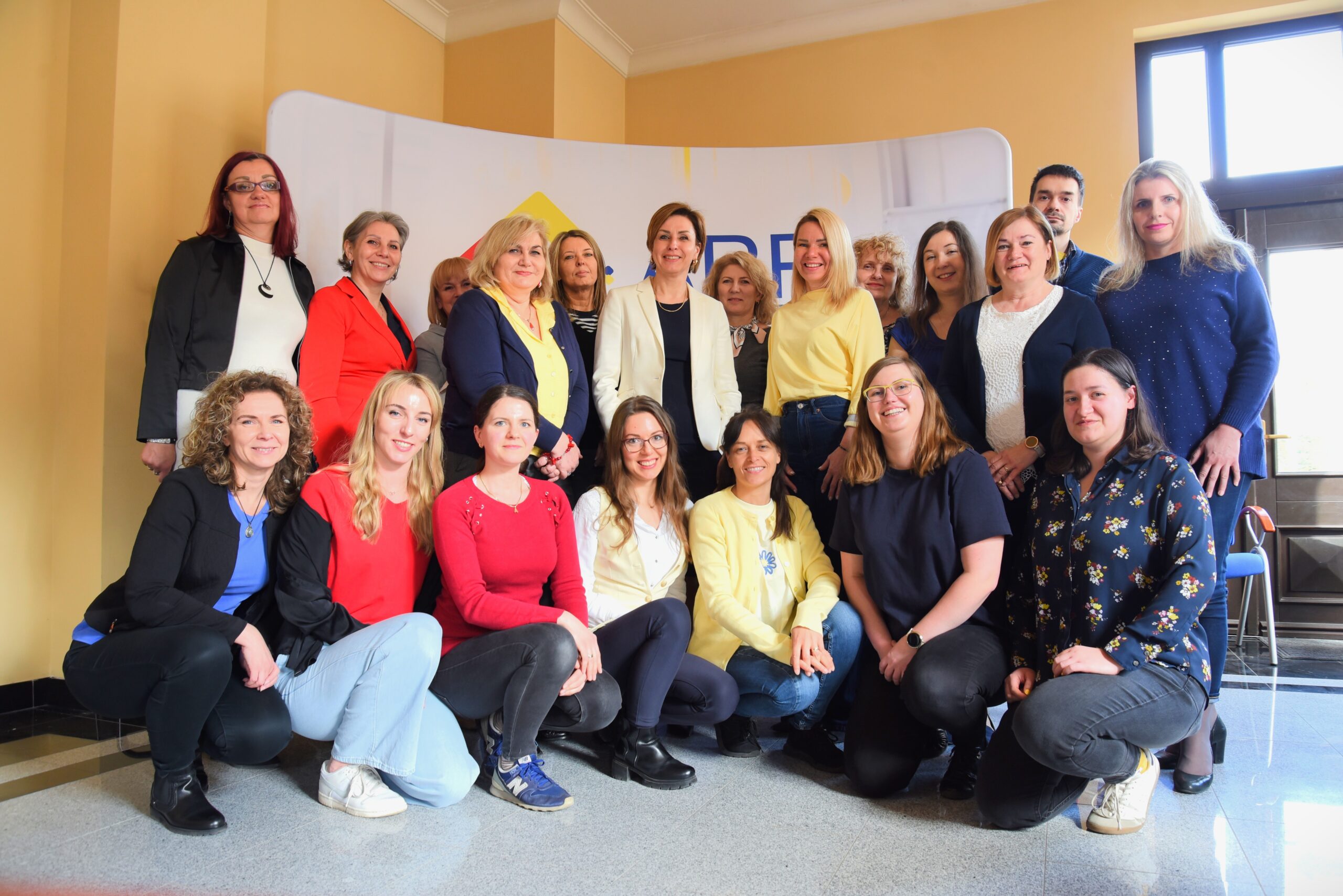 Grupa ludzi, głównie kobiet, ustawionych do zdjęcia grupowego, na tle żółtej ściany i białego baneru.